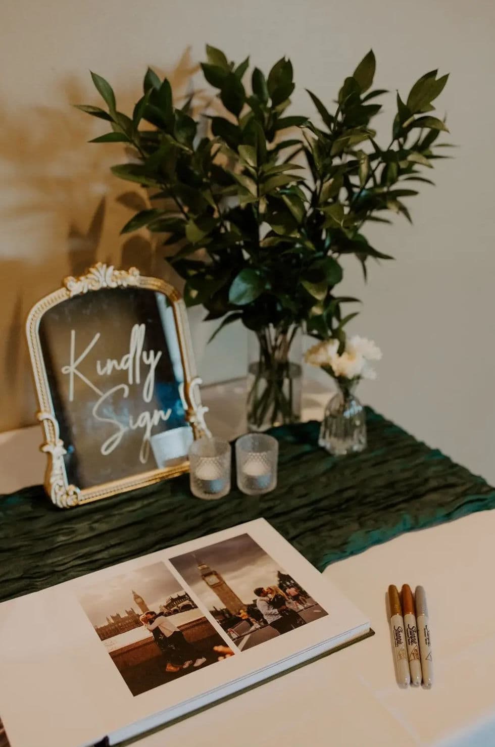Wedding guestbook setup with greenery, sign, and photos on a decorated table.