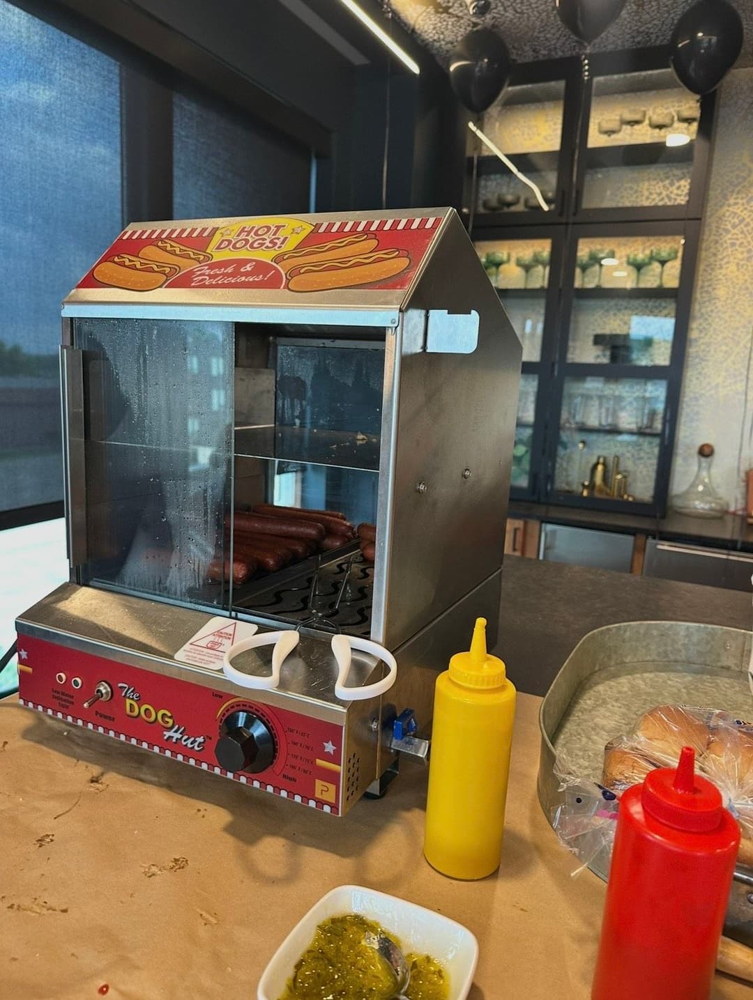 Hot dog cart with steaming hot dogs, condiments, and modern kitchen backdrop.