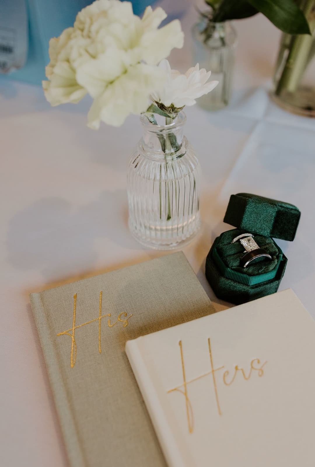 Hers and His wedding journals with flowers and a ring box on a table setting.