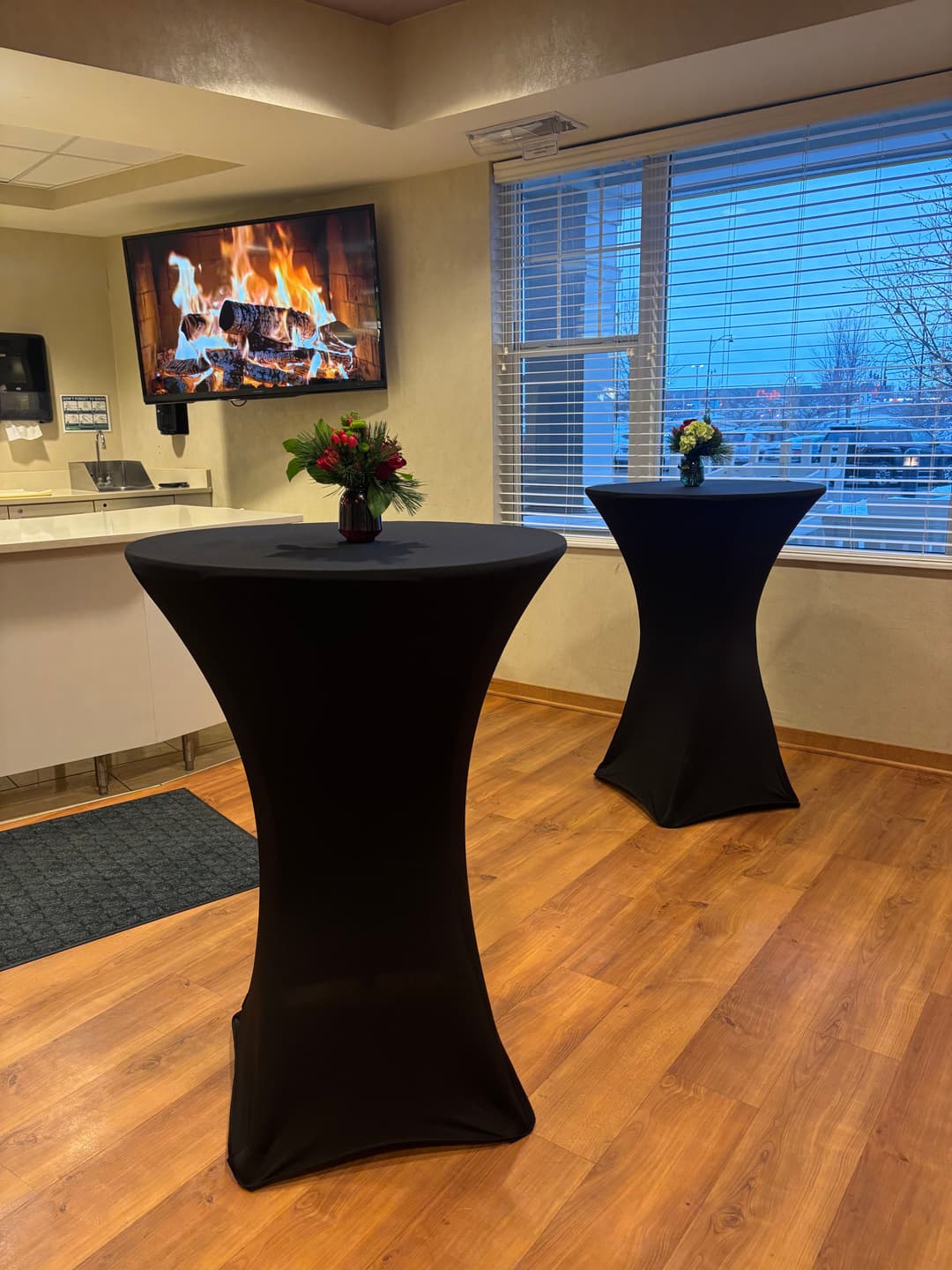 Two tall black cocktail tables with floral arrangements near a fireplace and large window.