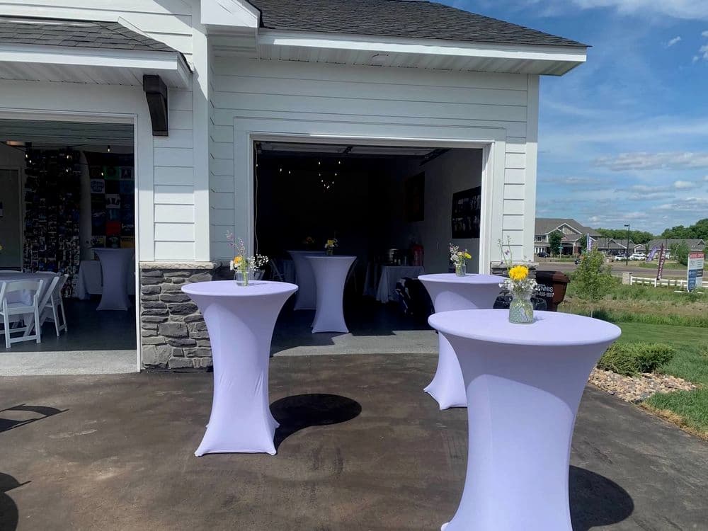 Elegant outdoor event with purple cocktail tables and floral centerpieces in a garage setting.